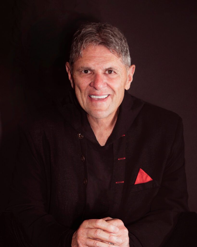 Smiling man in a black suit with a red pocket square against a dark background.