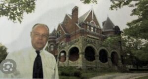 Man in front of a large, old brick mansion with arched windows and a steep roof.